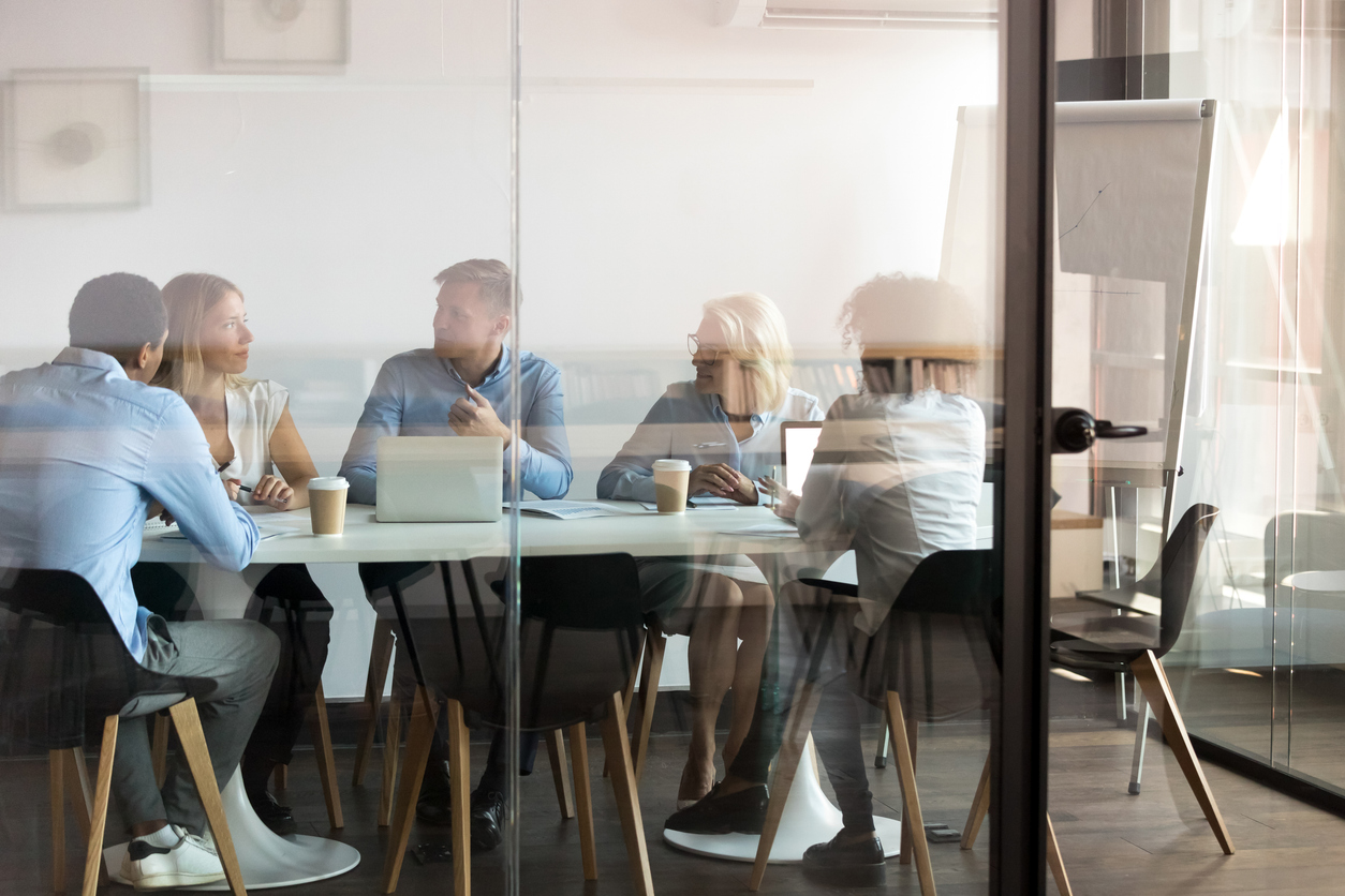 Advertising department brainstorming at modern office boardroom behind closed doors, view through the glass wall. Diverse staff discussing how to stretch an ad budget in a tough economy.